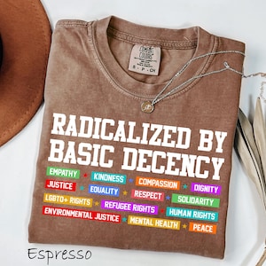 May include: Espresso-colored t-shirt with the text "RADICALIZED BY BASIC DECENCY" in white. Below the text are colorful labels with words like "EMPATHY", "JUSTICE", and "EQUALITY". A silver necklace with a pendant is on the shirt.