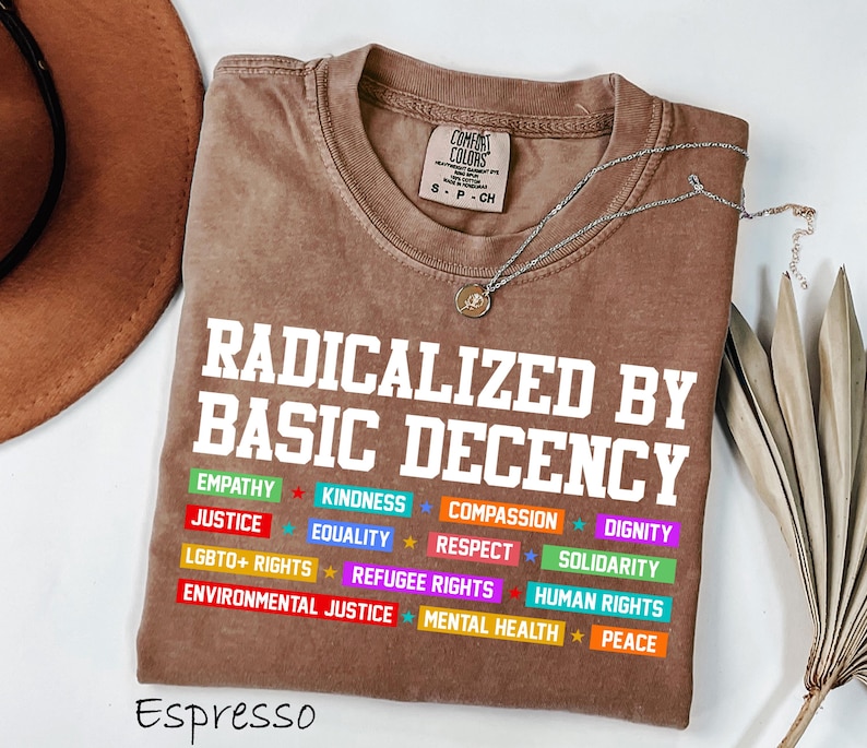 May include: Espresso-colored t-shirt with the text "RADICALIZED BY BASIC DECENCY" in white. Below the text are colorful labels with words like "EMPATHY", "JUSTICE", and "EQUALITY". A silver necklace with a pendant is on the shirt.