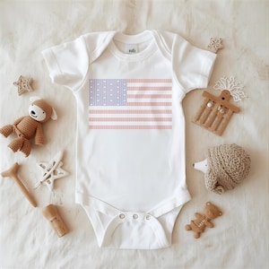 A white onesie with an American flag design is displayed on a white surface, surrounded by various baby items such as a teddy bear, a hedgehog, and a star.