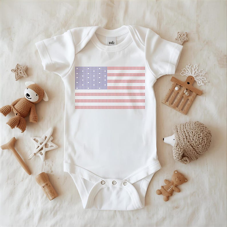 A white onesie with an American flag design is displayed on a white surface, surrounded by various baby items such as a teddy bear, a hedgehog, and a star.