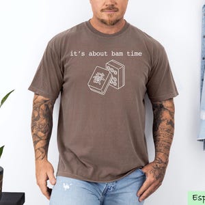 A man wearing a brown t-shirt with a graphic design and text printed on it, standing in front of a plant and a denim jacket.