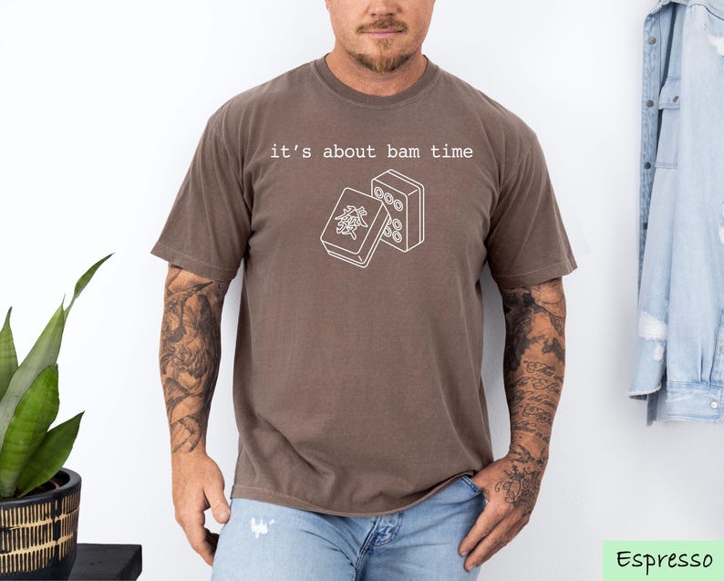 A man wearing a brown t-shirt with a graphic design and text printed on it, standing in front of a plant and a denim jacket.