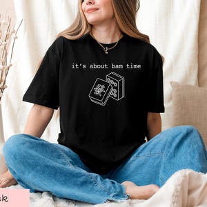 A woman wearing a black t-shirt with a graphic design and text, sitting on a bed with a vase of flowers and a pillow.