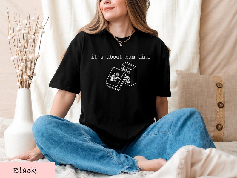 A woman wearing a black t-shirt with a graphic design and text, sitting on a bed with a vase of flowers and a pillow.