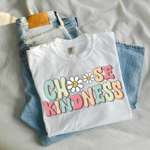 Puede incluir: Camiseta blanca con un gr&aacute;fico colorido que dice "Choose Kindness" con un dise&ntilde;o de margarita. La camiseta est&aacute; doblada y colocada encima de un par de pantalones vaqueros azules.