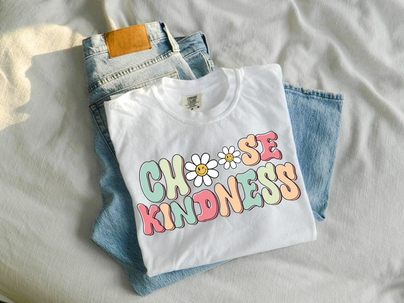 Puede incluir: Camiseta blanca con un gr&aacute;fico colorido que dice "Choose Kindness" con un dise&ntilde;o de margarita. La camiseta est&aacute; doblada y colocada encima de un par de pantalones vaqueros azules.