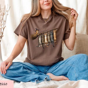 A woman sitting on a bed with a brown t-shirt featuring a graphic of cats on a broom.