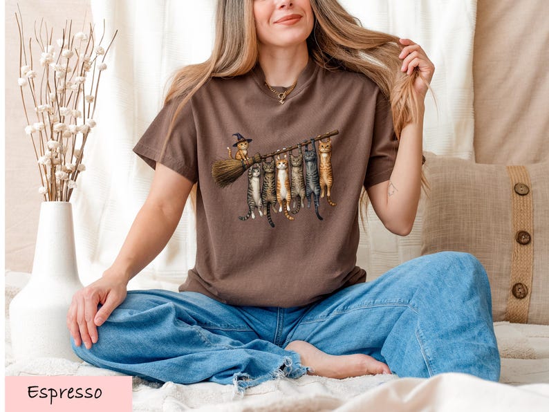 A woman sitting on a bed with a brown t-shirt featuring a graphic of cats on a broom.