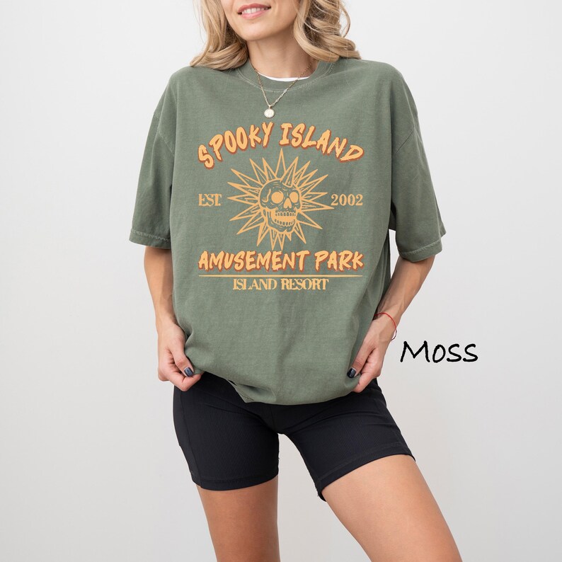 May include: Moss green t-shirt with the text "Spooky Island Amusement Park Island Resort" in orange. The design includes a skull and sunburst graphic. The shirt is paired with black shorts.