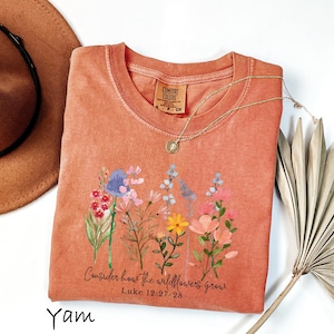May include: A coral-colored Comfort Colors t-shirt featuring a floral design with the text "Consider how the wildflowers grow" and the Bible verse Luke 12:27-28. A gold necklace and a brown hat are also in the image.