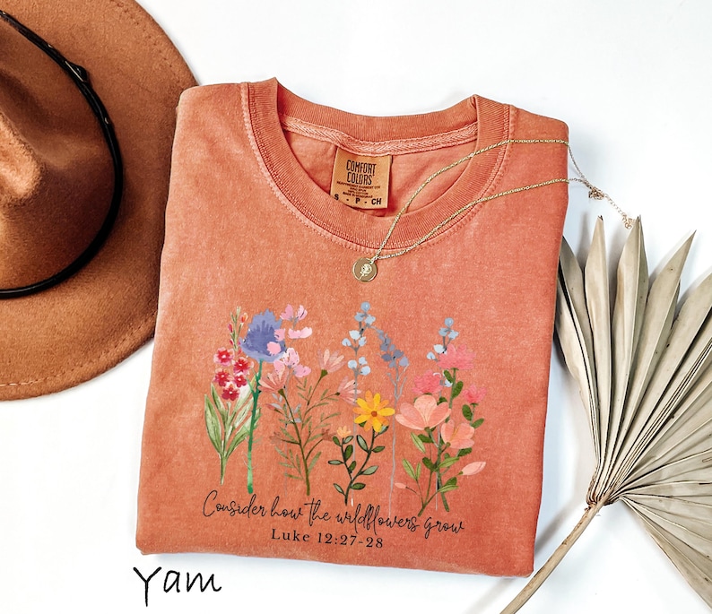 May include: A coral-colored Comfort Colors t-shirt featuring a floral design with the text "Consider how the wildflowers grow" and the Bible verse Luke 12:27-28. A gold necklace and a brown hat are also in the image.