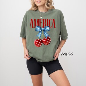 May include: A green t-shirt with the word "AMERICA" printed in red letters. The shirt features a red and white cherry with a blue and white star-patterned bow.