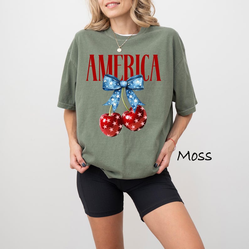 May include: A green t-shirt with the word "AMERICA" printed in red letters. The shirt features a red and white cherry with a blue and white star-patterned bow.