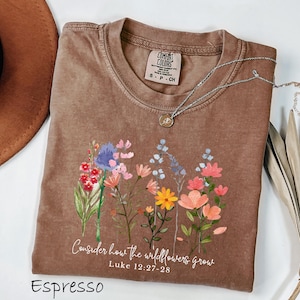 May include: Espresso-colored t-shirt with a floral design and the text "Consider how the wildflowers grow" from Luke 12:27-28. The shirt has a crew neck and a Comfort Colors tag. A silver necklace with a pendant is draped across the shirt.