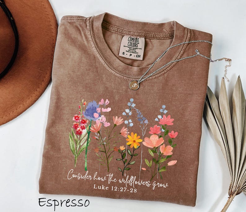 May include: Espresso-colored t-shirt with a floral design and the text "Consider how the wildflowers grow" from Luke 12:27-28. The shirt has a crew neck and a Comfort Colors tag. A silver necklace with a pendant is draped across the shirt.