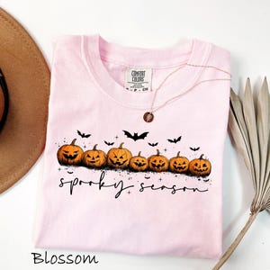 May include: A light pink t-shirt with a crew neck featuring a row of carved orange pumpkins and black bats. The words "spooky season" are written in a script font. The shirt is labeled "Blossom".