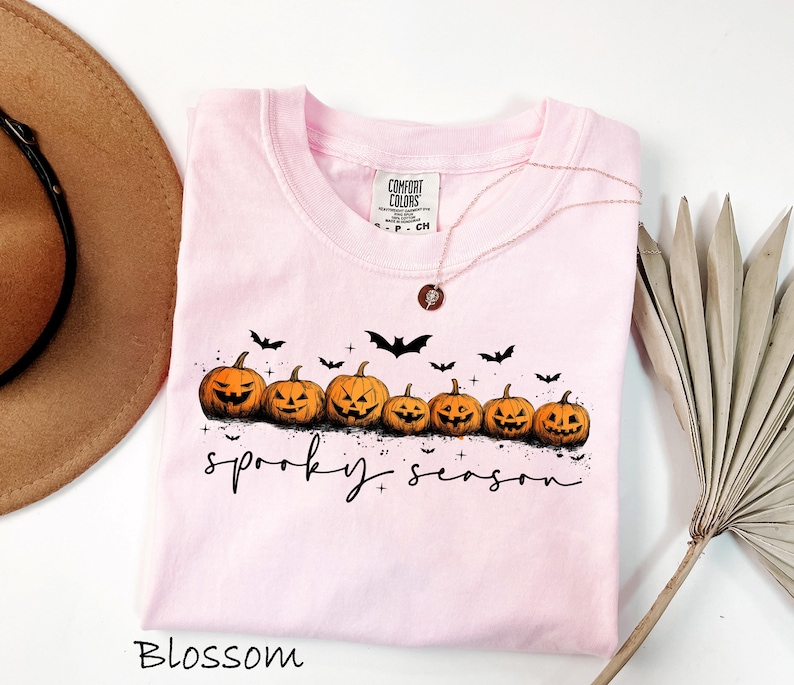 May include: A light pink t-shirt with a crew neck featuring a row of carved orange pumpkins and black bats. The words "spooky season" are written in a script font. The shirt is labeled "Blossom".