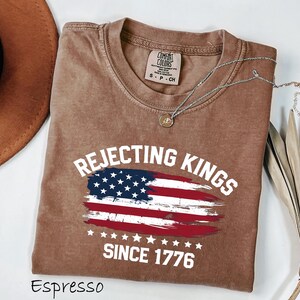May include: Espresso-colored t-shirt with the text "Rejecting Kings Since 1776" and an American flag design. The shirt has a crew neck and short sleeves. A silver necklace with a pendant is draped on the shirt.