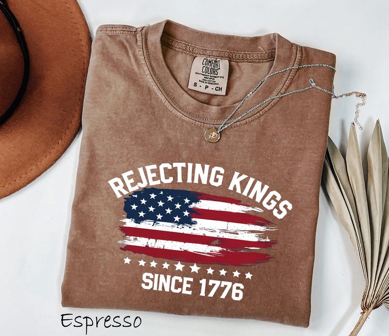 May include: Espresso-colored t-shirt with the text "Rejecting Kings Since 1776" and an American flag design. The shirt has a crew neck and short sleeves. A silver necklace with a pendant is draped on the shirt.