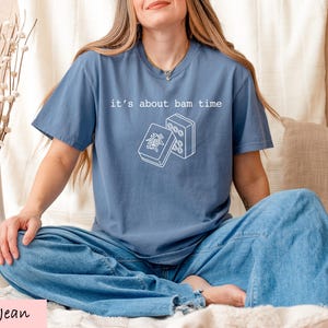 A woman wearing a blue t-shirt with a graphic design and text, sitting on a bed with a vase of flowers and a pillow.