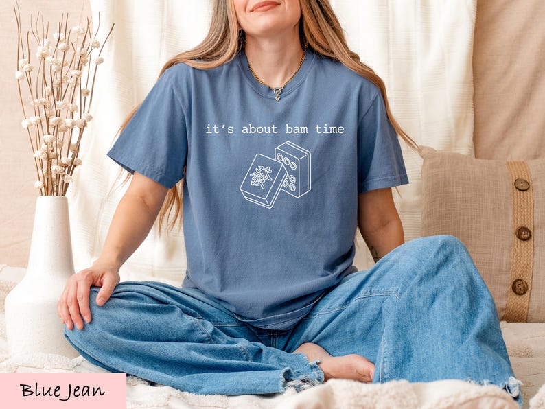 A woman wearing a blue t-shirt with a graphic design and text, sitting on a bed with a vase of flowers and a pillow.