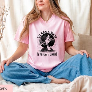 A woman wearing a pink t-shirt with a graphic design sits cross-legged on a bed, smiling.