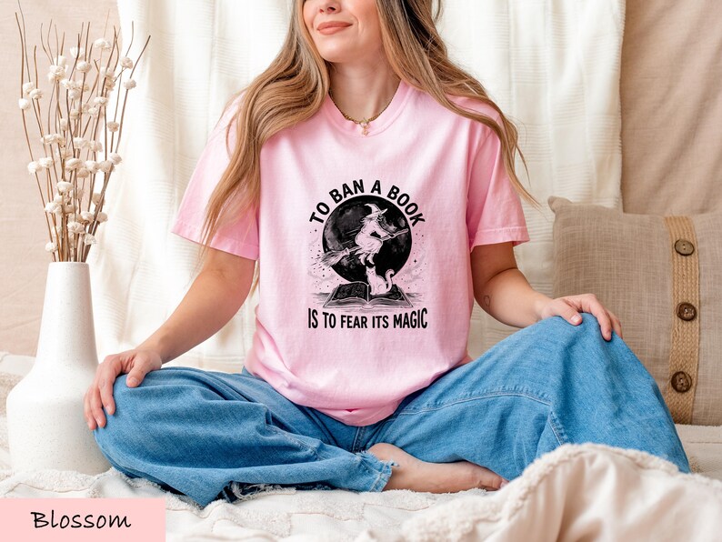 A woman wearing a pink t-shirt with a graphic design sits cross-legged on a bed, smiling.