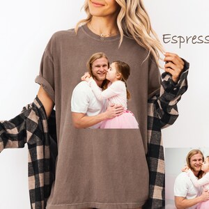 May include: Espresso-colored t-shirt featuring a printed image of a man and a young girl embracing. The woman in the image is wearing the t-shirt and a black and brown plaid shirt. The text "Espresso" is written on the top right of the image.