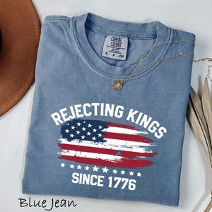 May include: Blue jean colored t-shirt with the text "Rejecting Kings Since 1776" and a distressed American flag design. The shirt has a crew neck and short sleeves. A gold necklace is draped across the shirt.