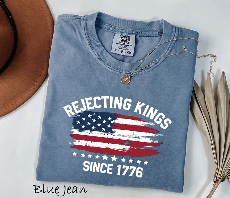May include: Blue jean colored t-shirt with the text "Rejecting Kings Since 1776" and a distressed American flag design. The shirt has a crew neck and short sleeves. A gold necklace is draped across the shirt.
