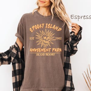 May include: Espresso-colored t-shirt with a distressed graphic that reads "Spooky Island Amusement Park Island Resort" with a skull and sunburst design. The shirt is worn with a black and brown plaid flannel.