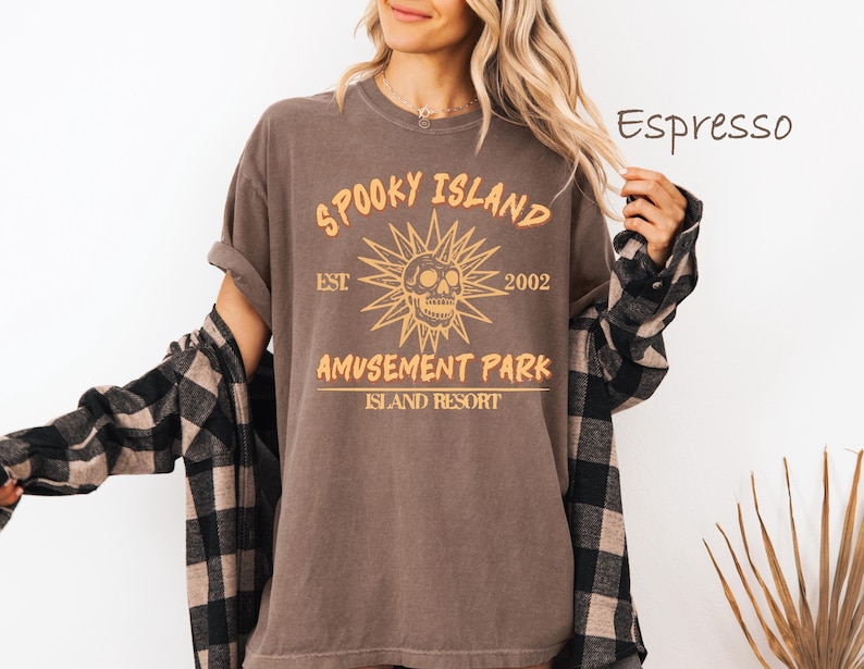 May include: Espresso-colored t-shirt with a distressed graphic that reads "Spooky Island Amusement Park Island Resort" with a skull and sunburst design. The shirt is worn with a black and brown plaid flannel.