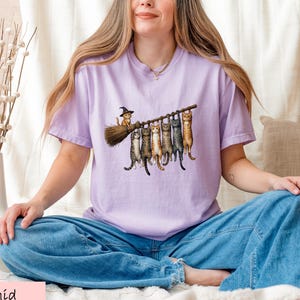A woman wearing a purple t-shirt with a graphic of cats sitting on a branch and holding a broom is sitting cross-legged on a bed.