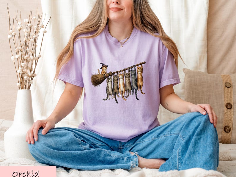 A woman wearing a purple t-shirt with a graphic of cats sitting on a branch and holding a broom is sitting cross-legged on a bed.