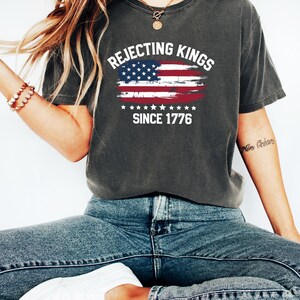 May include: Dark gray t-shirt with the text "Rejecting Kings Since 1776" and an American flag design. The shirt is a comfortable, casual style.