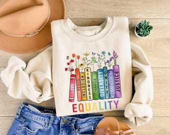 May include: A cream-coloured crewneck sweatshirt featuring a graphic of colourful books standing upright, each labelled with words like Love, Peace, Inclusion, Kindness, Change, Diversity, Unity, Compassion, and Justice, topped with various wildflowers.