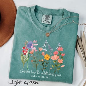 May include: Light green t-shirt with a floral design and the text "Consider how the wildflowers grow" from Luke 12:27-28. The shirt has a crew neck and short sleeves. A silver necklace with a pendant is draped across the shirt.
