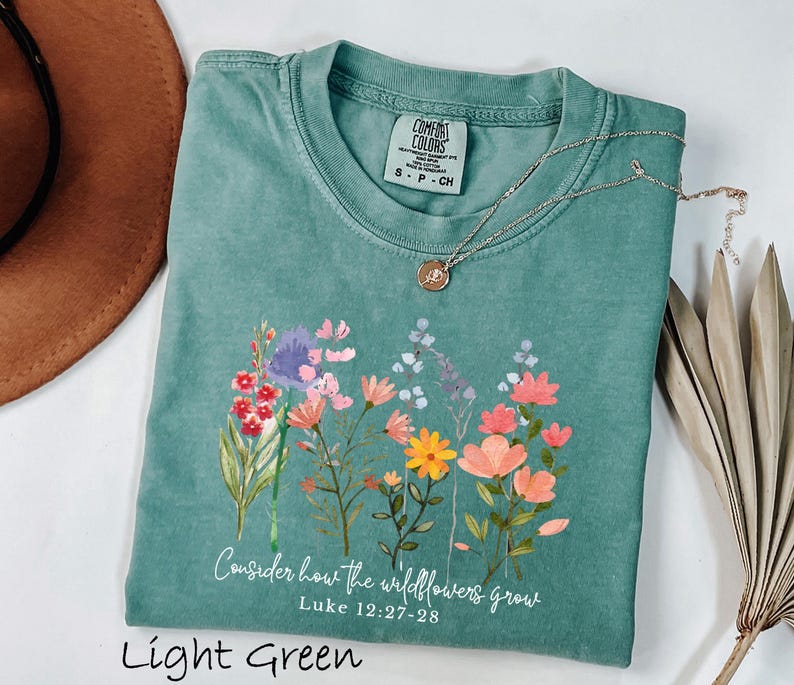 May include: Light green t-shirt with a floral design and the text "Consider how the wildflowers grow" from Luke 12:27-28. The shirt has a crew neck and short sleeves. A silver necklace with a pendant is draped across the shirt.