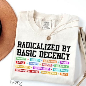 May include: Ivory-colored t-shirt with the text "RADICALIZED BY BASIC DECENCY" in black. Below, colorful text blocks list values like "EMPATHY," "JUSTICE," and "EQUALITY." A gold necklace and a brown hat are also visible.