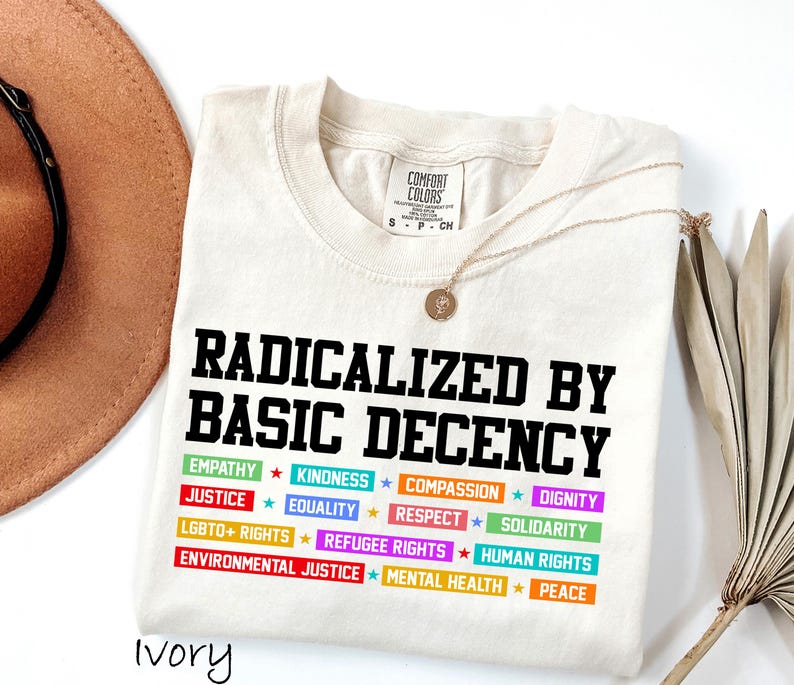 May include: Ivory-colored t-shirt with the text "RADICALIZED BY BASIC DECENCY" in black. Below, colorful text blocks list values like "EMPATHY," "JUSTICE," and "EQUALITY." A gold necklace and a brown hat are also visible.