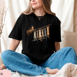 A woman wearing a black t-shirt with a graphic of cats sitting on a broom, smiling and looking at the camera.
