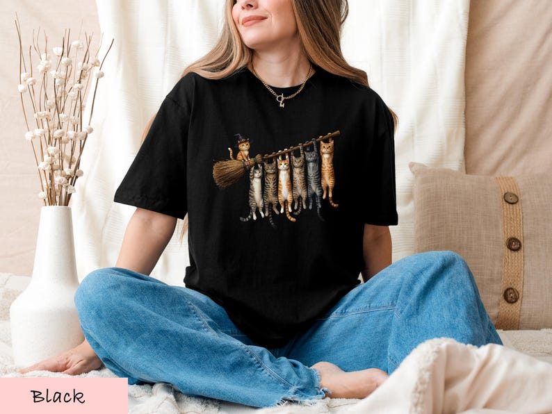 A woman wearing a black t-shirt with a graphic of cats sitting on a broom, smiling and looking at the camera.