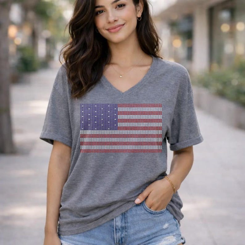 A young woman with long brown hair is standing outdoors, wearing a gray t-shirt with an American flag design. She is smiling and posing for the camera.