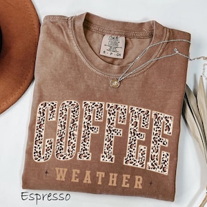 May include: Espresso-coloured t-shirt featuring the words "COFFEE WEATHER" in a leopard print design. The shirt has a crew neck and short sleeves. A silver necklace with a small pendant is draped across the shirt.