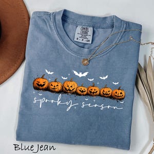 May include: Blue jean colored t-shirt with a row of carved orange pumpkins and white bats. The words "spooky season" are written in white script. A gold necklace is draped across the shirt.