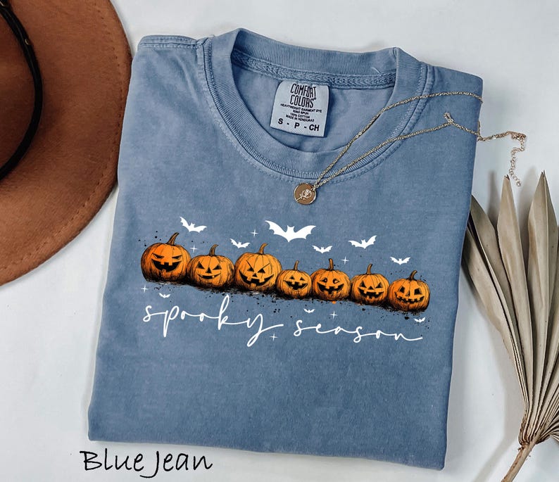 May include: Blue jean colored t-shirt with a row of carved orange pumpkins and white bats. The words "spooky season" are written in white script. A gold necklace is draped across the shirt.