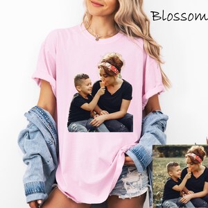 May include: A light pink t-shirt featuring a photograph of a mother and child sharing an ice cream cone. The child is offering the ice cream to the mother. The word "Blossom" is written in cursive in the upper right corner. The model is wearing a denim jacket.