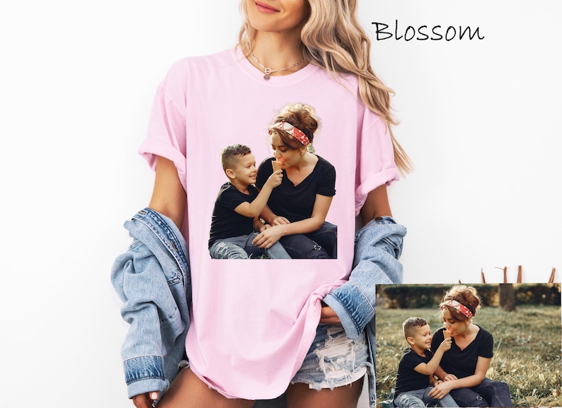 May include: A light pink t-shirt featuring a photograph of a mother and child sharing an ice cream cone. The child is offering the ice cream to the mother. The word "Blossom" is written in cursive in the upper right corner. The model is wearing a denim jacket.