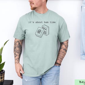 A man wearing a light green t-shirt with a graphic design and text printed on it, standing in front of a plant and a denim jacket.