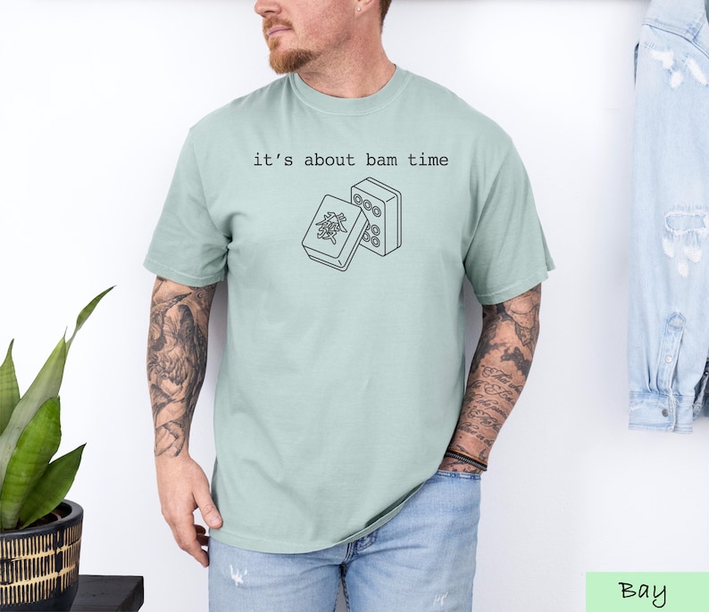 A man wearing a light green t-shirt with a graphic design and text printed on it, standing in front of a plant and a denim jacket.